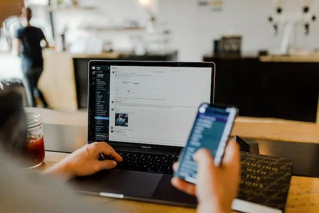 A person is seated at a wooden table using Slack in a laptop and holding a smartphone